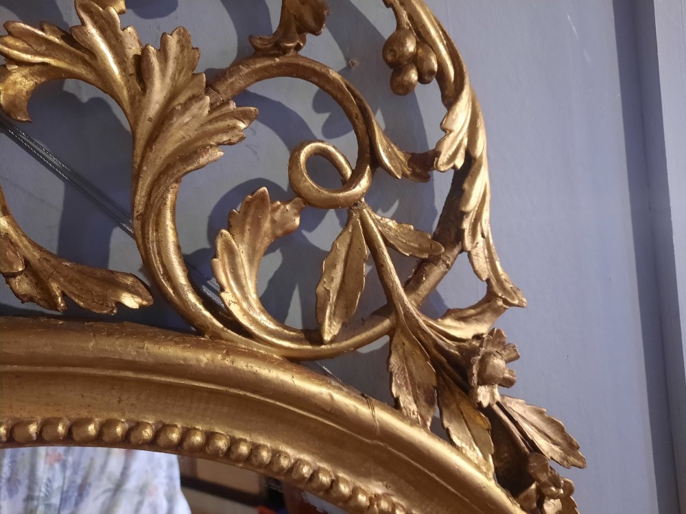 An oval giltwood wall mirror featuring an intricate carved urn cresting, acanthus leaf foliage, and a beaded inner frame border. View VI
