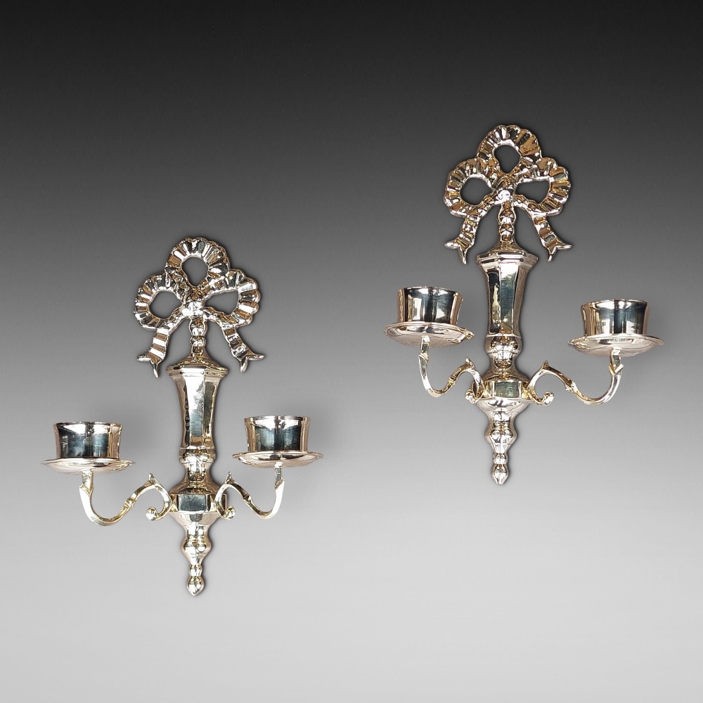 A pair of polished brass double-arm wall sconces featuring ribbon bow top motifs, urn-shaped central stems, and scrolling candle branches.