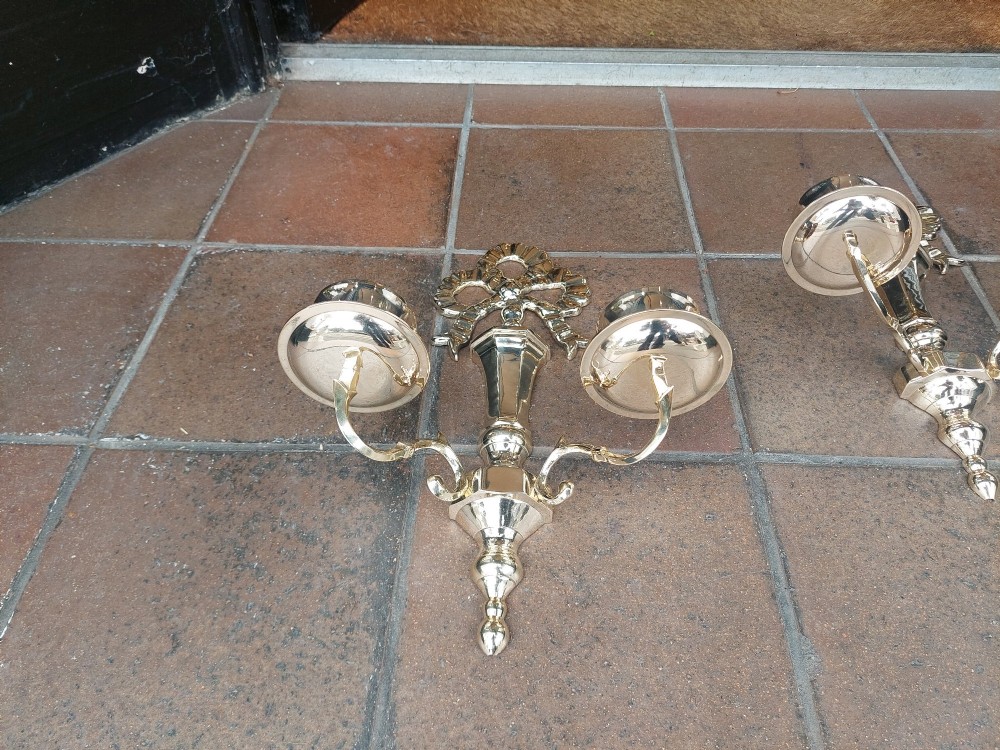 A pair of polished brass double-arm wall sconces featuring ribbon bow top motifs, urn-shaped central stems, and scrolling candle branches. View V