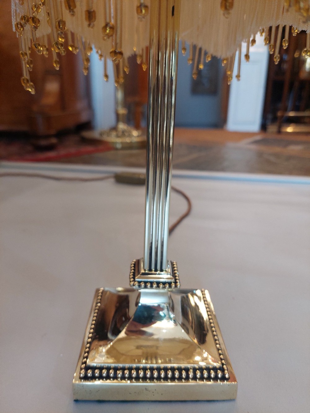 An Edwardian Neoclassical Revival silver-plated table lamp featuring a fluted column stem, square stepped base, and a cream fabric shade adorned with beaded fringe. View VI