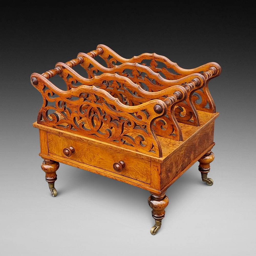 A High Victorian burr walnut Canterbury featuring four open compartments with fretwork dividers above a single drawer with turned legs on brass castors.