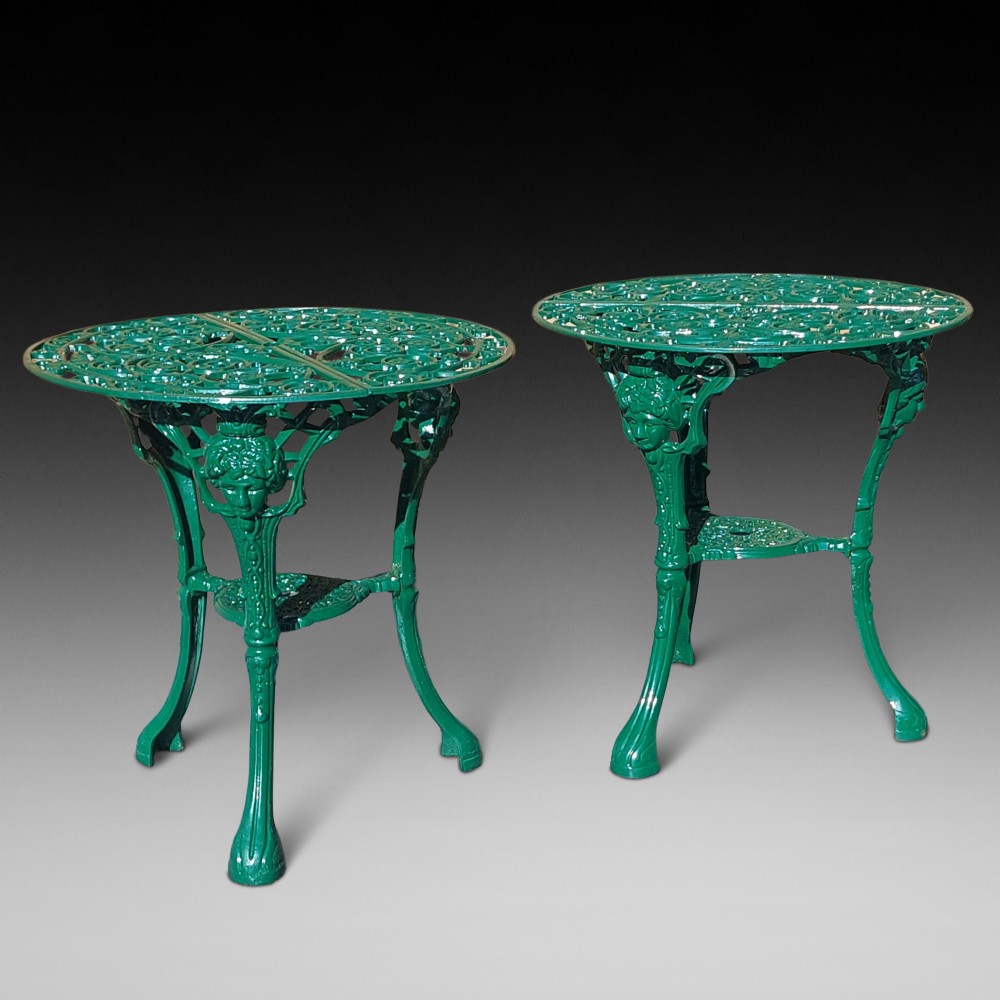 A pair of circular green painted cast iron garden tables featuring pierced foliate tops, mask-adorned legs, and a lower triangular stretcher shelf.