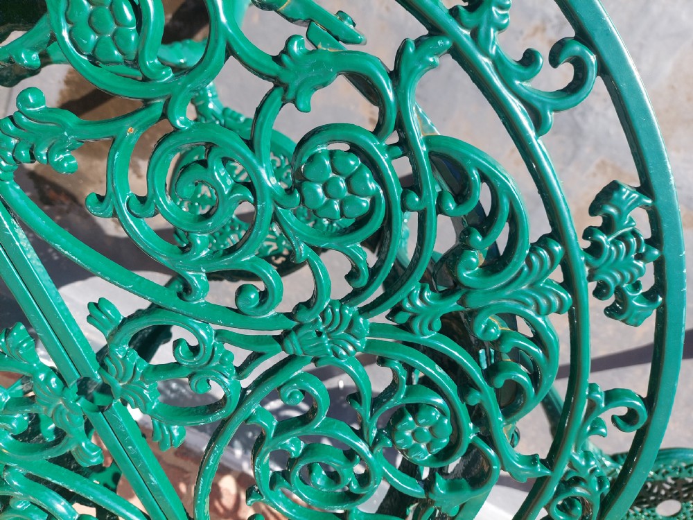 A pair of circular green painted cast iron garden tables featuring pierced foliate tops, mask-adorned legs, and a lower triangular stretcher shelf. View IV