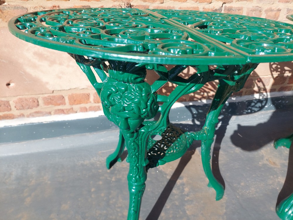 A pair of circular green painted cast iron garden tables featuring pierced foliate tops, mask-adorned legs, and a lower triangular stretcher shelf. View V