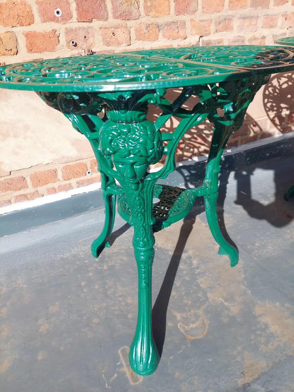 A pair of circular green painted cast iron garden tables featuring pierced foliate tops, mask-adorned legs, and a lower triangular stretcher shelf. View VII