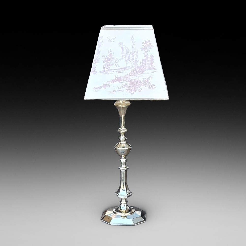 An Art Nouveau silvered brass table lamp featuring a hexagonal faceted stem and a white tapered shade printed with a pastoral scene.