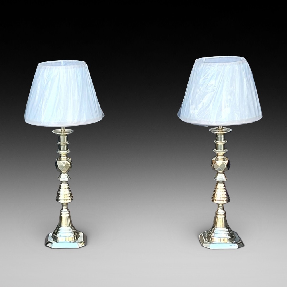 A pair of turned brass Edwardian candlestick table lamps fitted with white pleated lampshades.