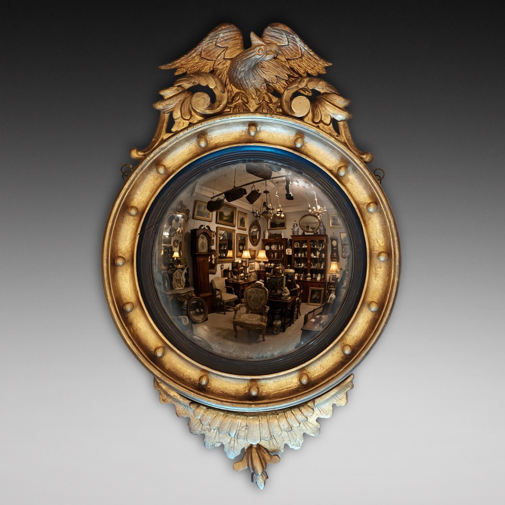 A circular Regency giltwood convex wall mirror featuring a carved eagle surmount, spherical balls around the moulded frame, a black reeded slip, and an acanthus leaf apron.