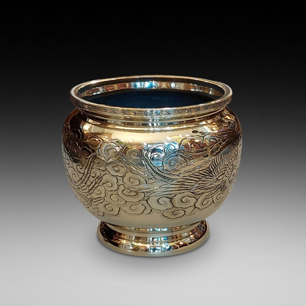 A rounded polished brass Japanese jardiniere with hand-chased relief dragons and clouds, resting on a stepped base.