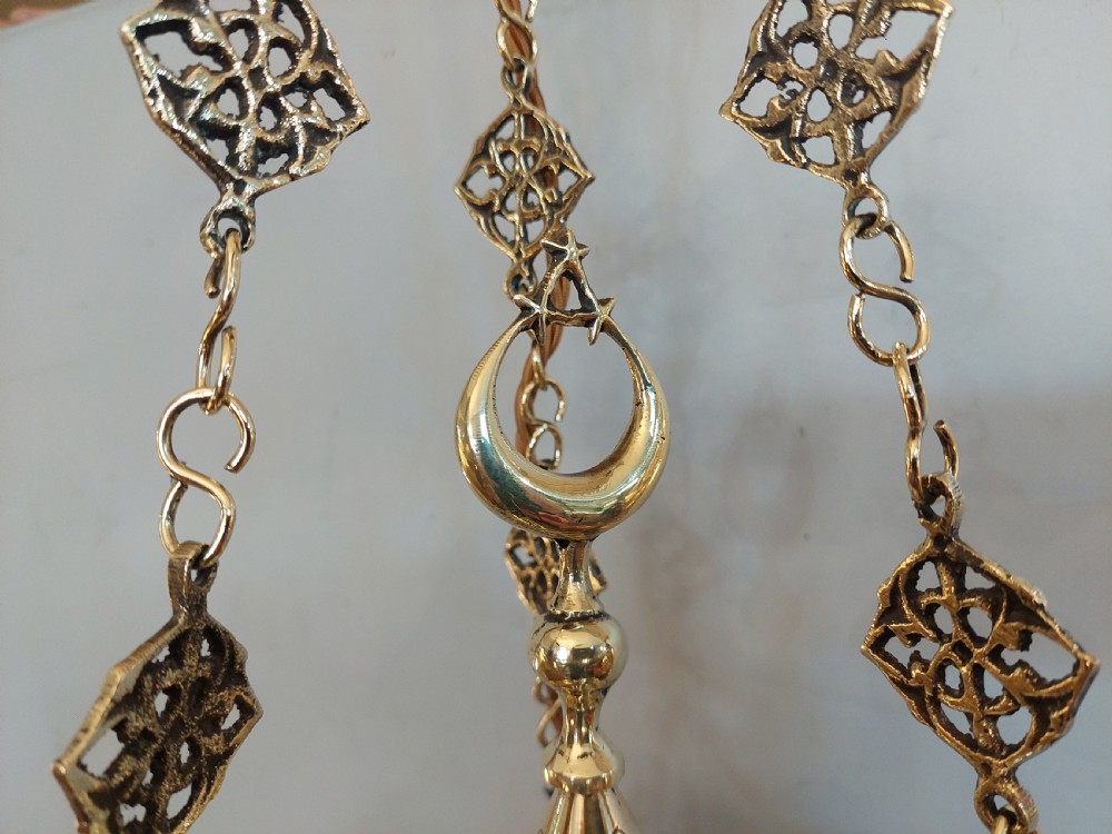A hanging polished brass lantern featuring intricate pierced fretwork, dark glass insert panels, and three suspension chains topped with a crescent moon motif. View IV