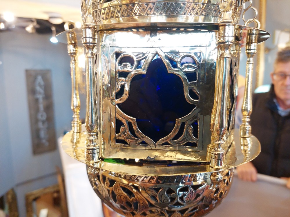 A hanging polished brass lantern featuring intricate pierced fretwork, dark glass insert panels, and three suspension chains topped with a crescent moon motif. View VI
