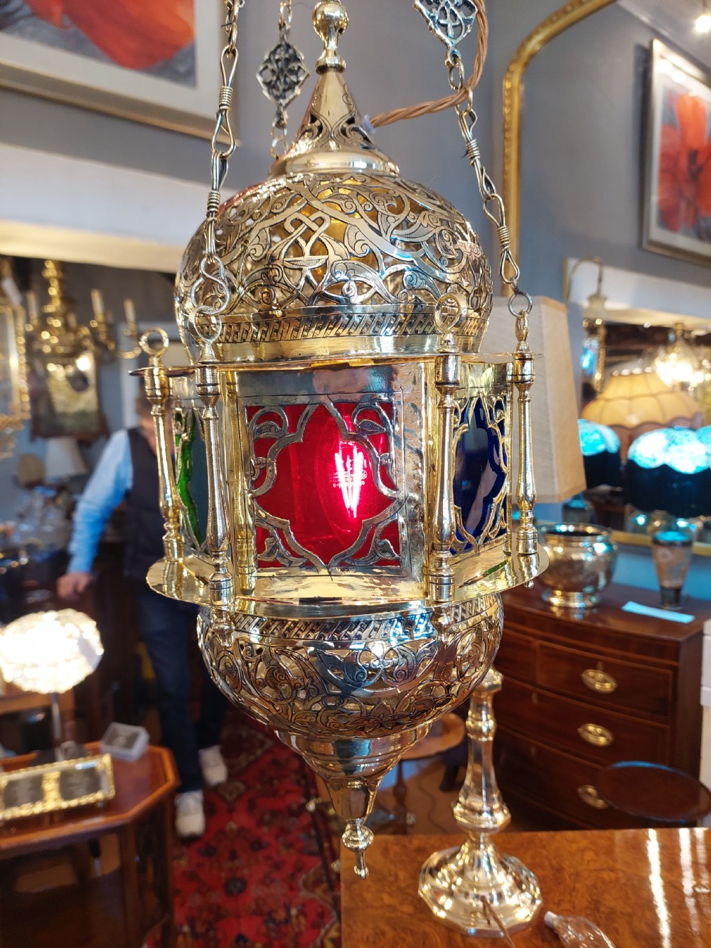 A hanging polished brass lantern featuring intricate pierced fretwork, dark glass insert panels, and three suspension chains topped with a crescent moon motif. View X