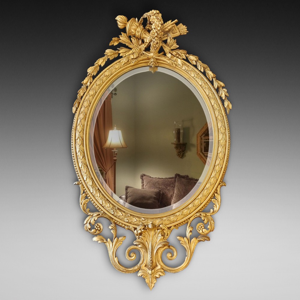 An oval giltwood wall mirror with carved cresting featuring a torch, quiver of arrows, and leaf garlands above an acanthus scroll base.