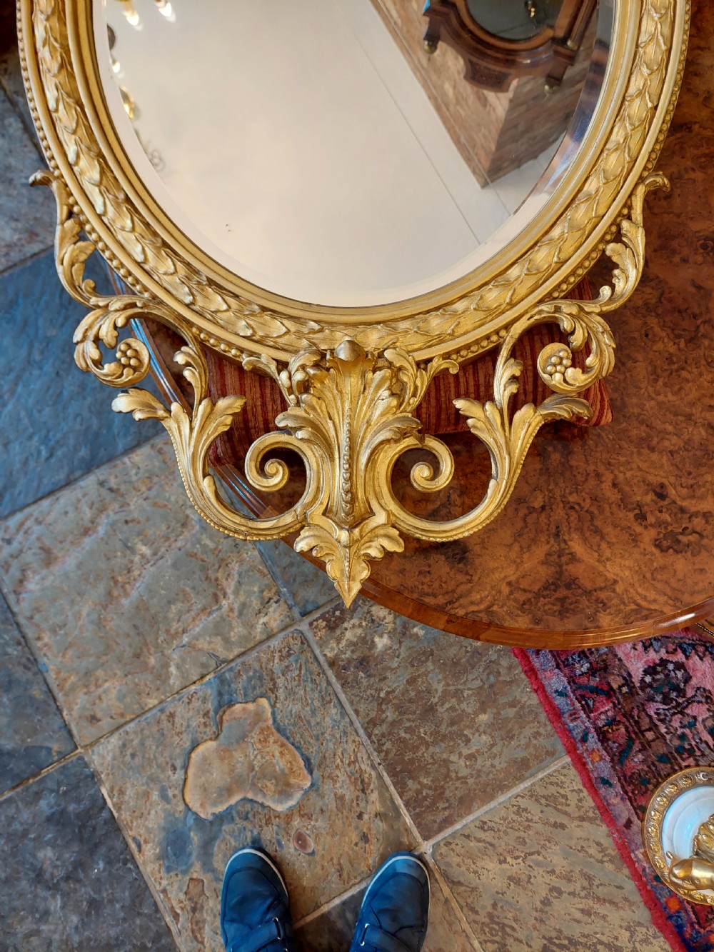 An oval giltwood wall mirror with carved cresting featuring a torch, quiver of arrows, and leaf garlands above an acanthus scroll base. View II