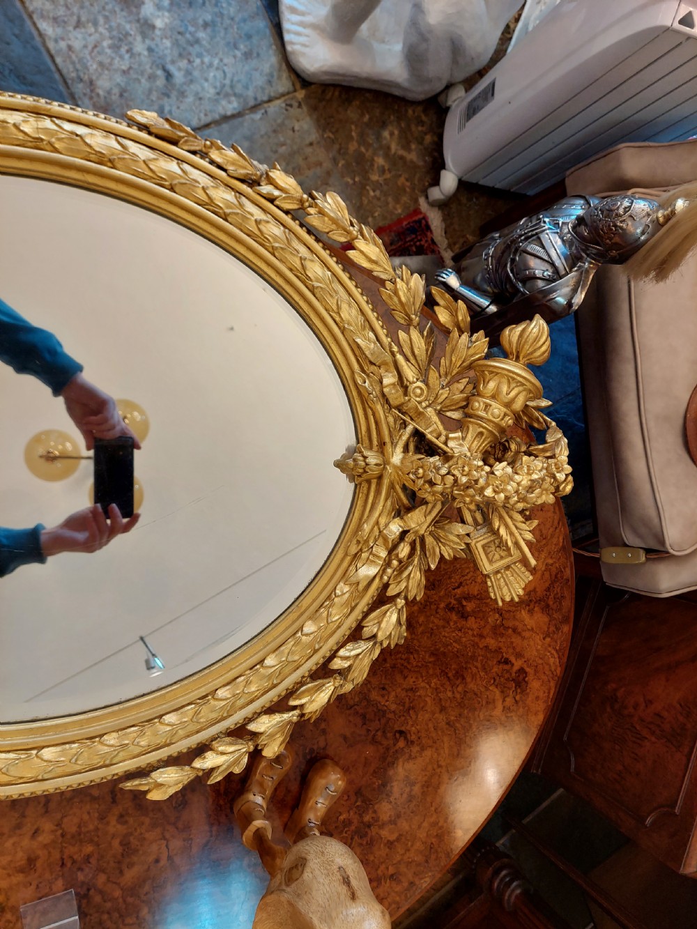 An oval giltwood wall mirror with carved cresting featuring a torch, quiver of arrows, and leaf garlands above an acanthus scroll base. View III