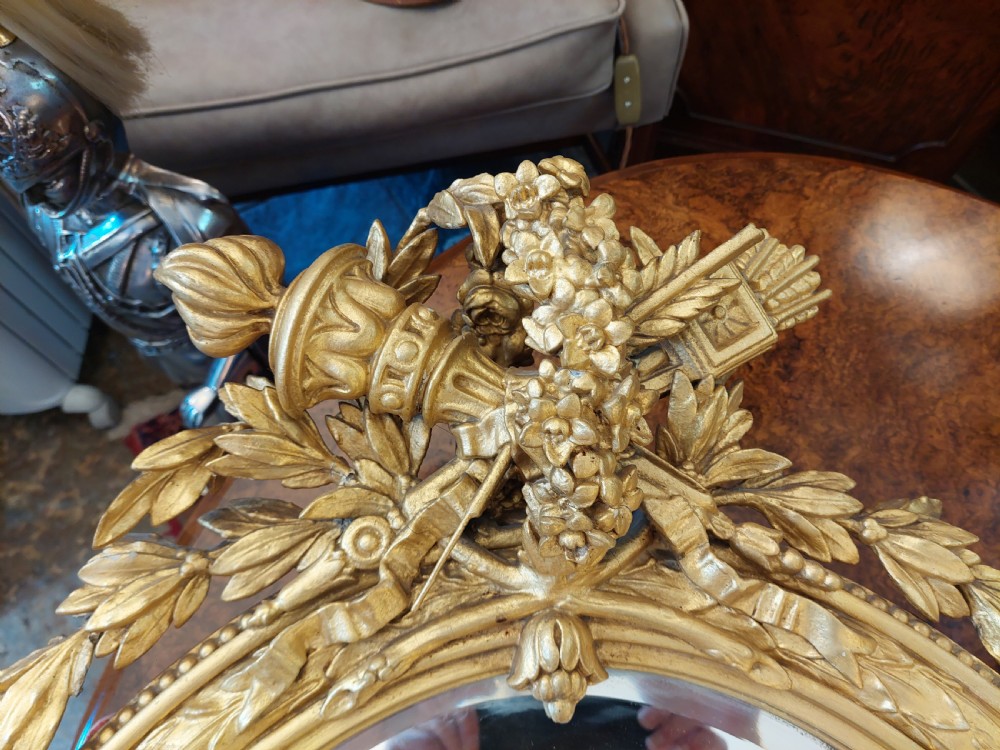 An oval giltwood wall mirror with carved cresting featuring a torch, quiver of arrows, and leaf garlands above an acanthus scroll base. View IV