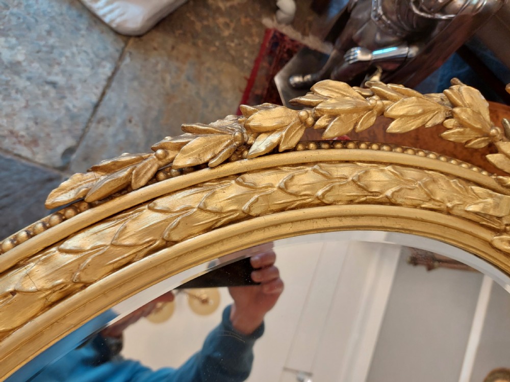 An oval giltwood wall mirror with carved cresting featuring a torch, quiver of arrows, and leaf garlands above an acanthus scroll base. View V