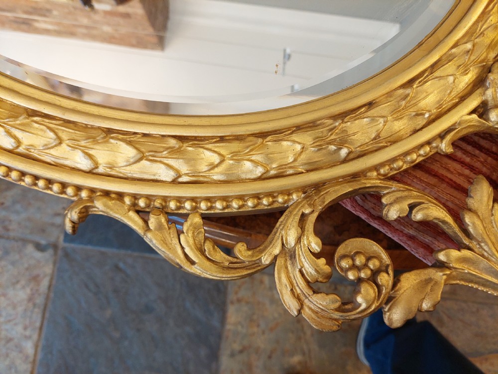 An oval giltwood wall mirror with carved cresting featuring a torch, quiver of arrows, and leaf garlands above an acanthus scroll base. View VI