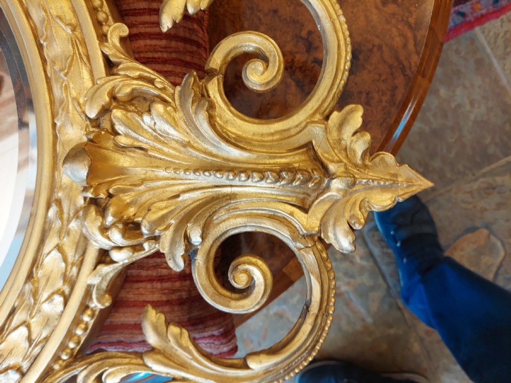An oval giltwood wall mirror with carved cresting featuring a torch, quiver of arrows, and leaf garlands above an acanthus scroll base. View VII