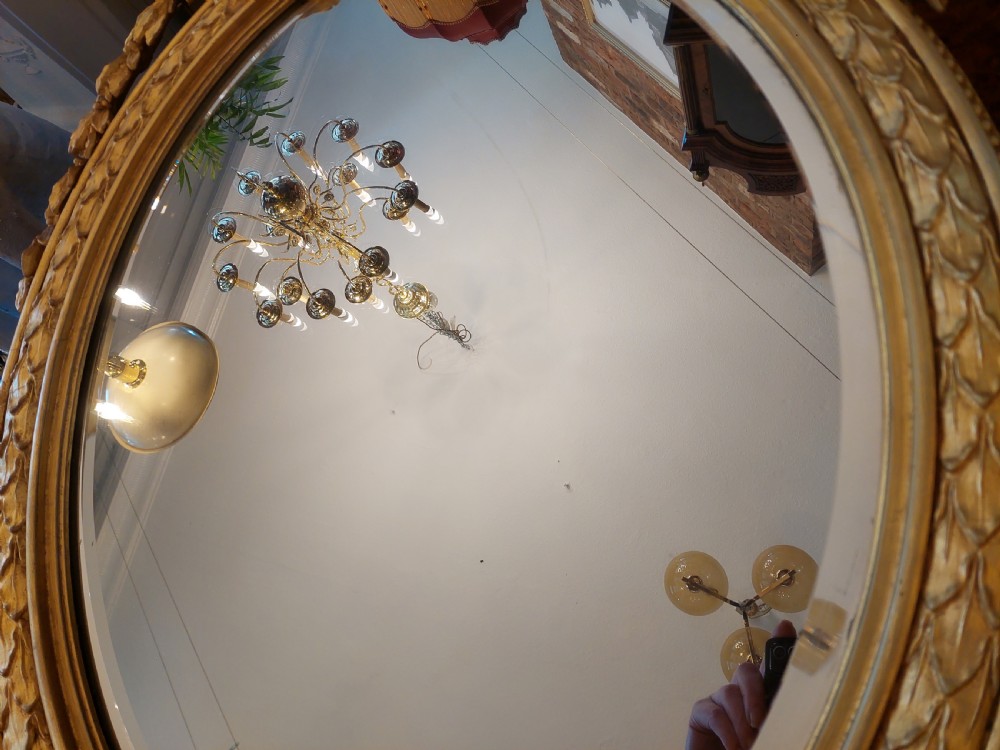 An oval giltwood wall mirror with carved cresting featuring a torch, quiver of arrows, and leaf garlands above an acanthus scroll base. View IX