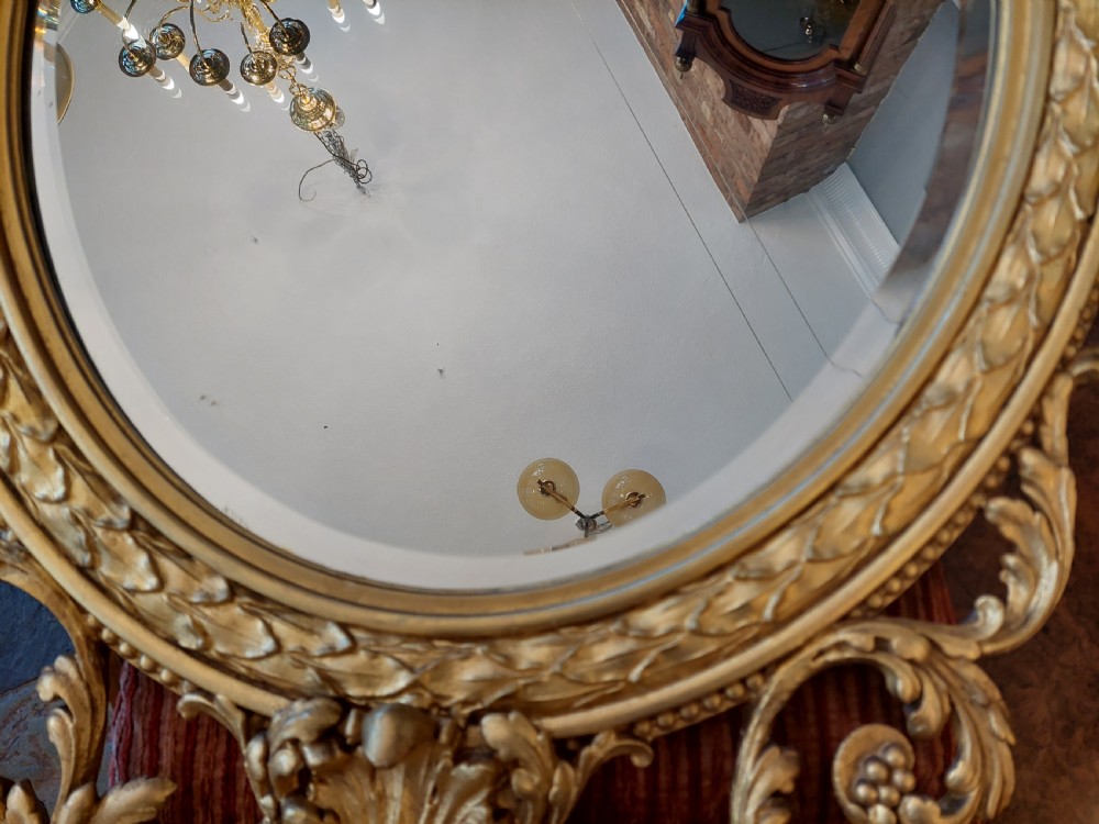 An oval giltwood wall mirror with carved cresting featuring a torch, quiver of arrows, and leaf garlands above an acanthus scroll base. View X