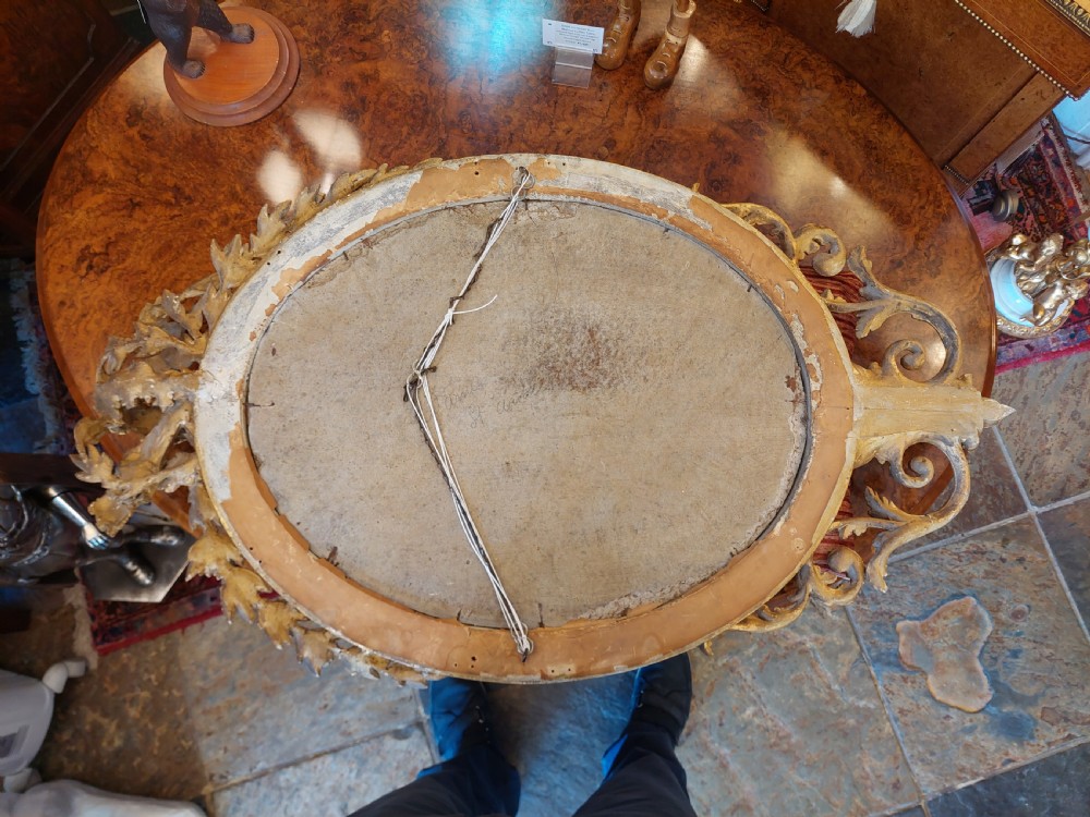 An oval giltwood wall mirror with carved cresting featuring a torch, quiver of arrows, and leaf garlands above an acanthus scroll base. View XI