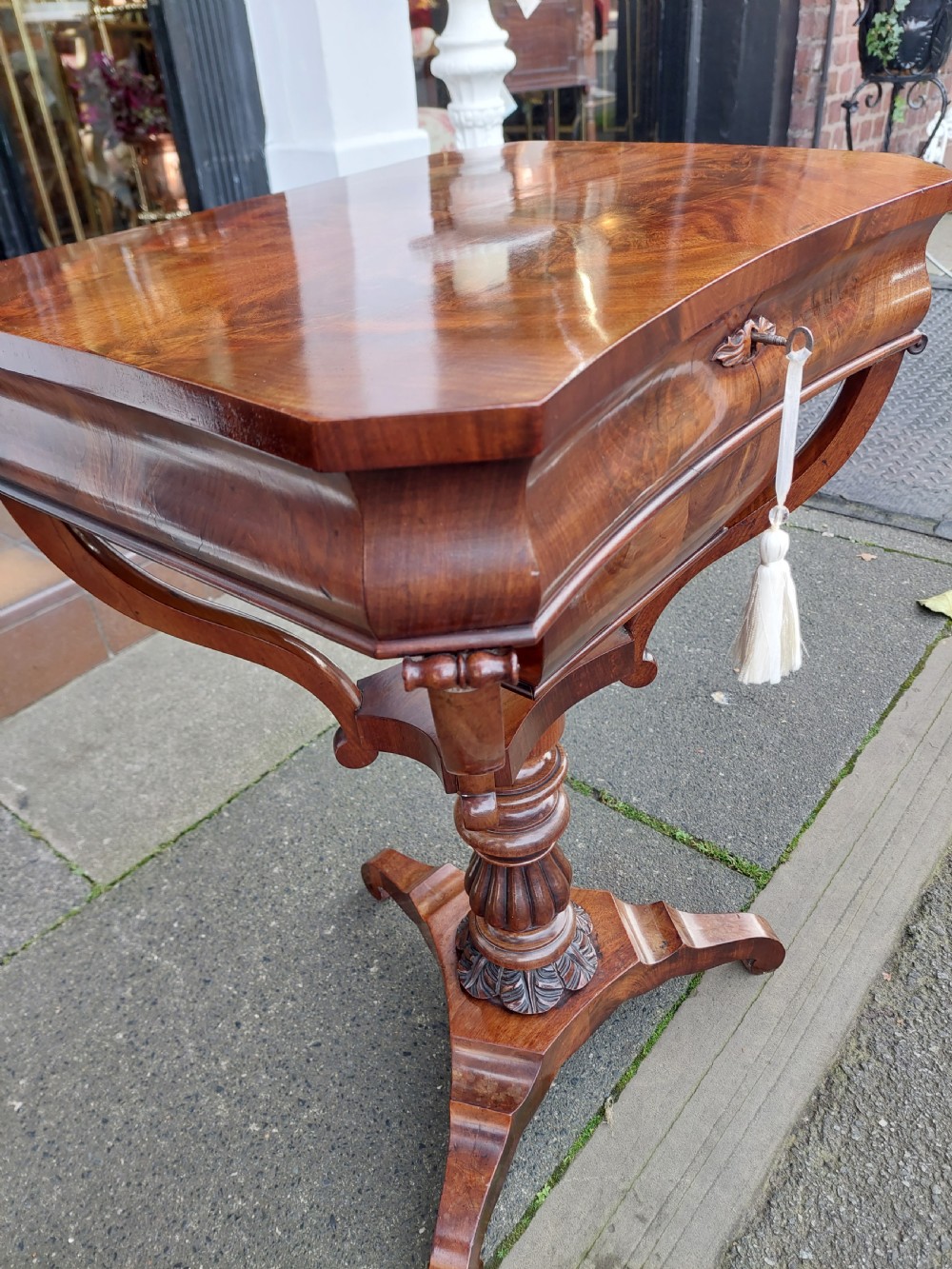 A flame mahogany Victorian sewing table featuring a canted corner top, a faceted turned pedestal column, and a scroll-cut tripod base. View VI