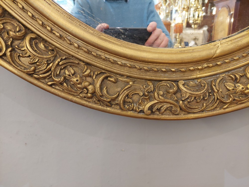A nineteenth-century oval wall mirror featuring a heavily carved giltwood frame with floral and scrolling moulding details. View II