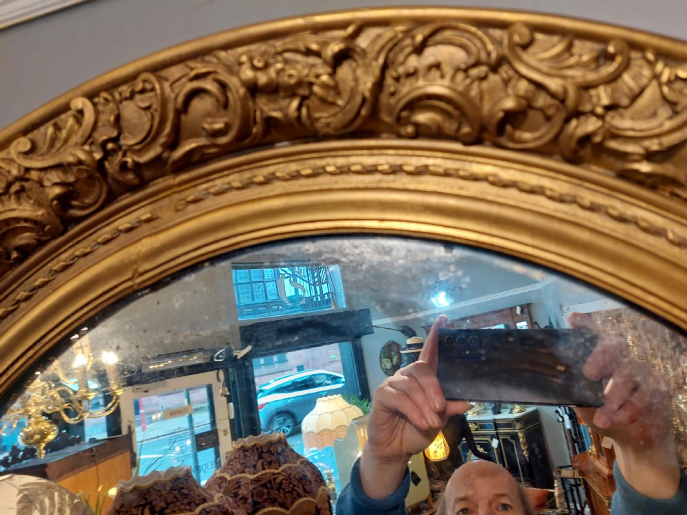 A nineteenth-century oval wall mirror featuring a heavily carved giltwood frame with floral and scrolling moulding details. View VI