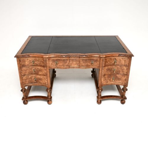 fig 1. An antique burr walnut partners desk featuring a black leather writing surface, multiple serpentine front drawers with brass handles, and turned legs joined by stretchers.