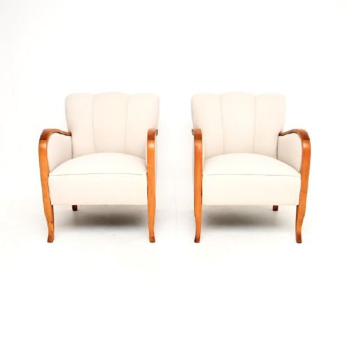 fig 1. A pair of Swedish Art Deco armchairs featuring off-white fabric upholstery, fluted backrests, and curved satin birch armrests extending down to tapered legs.