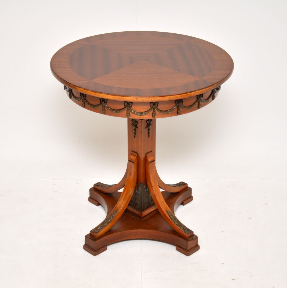 A circular antique mahogany occasional table featuring a segmented veneered top, metal festoon mounts along the apron, and a central pedestal with outcurved legs.