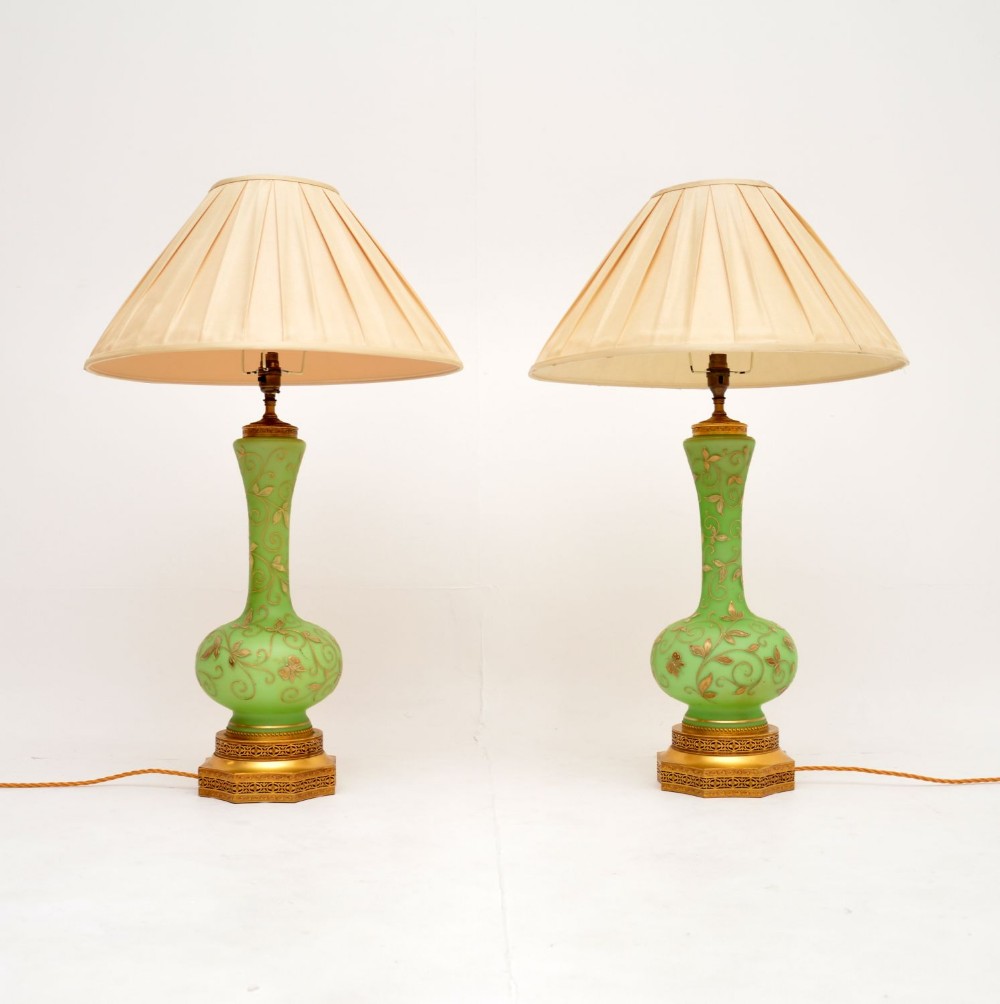 A pair of antique green glass table lamps decorated with gilt floral motifs, raised on ornate gold bases with pleated cream shades.