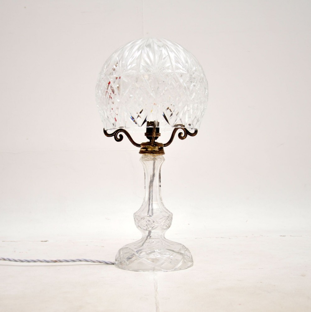 A clear cut crystal table lamp featuring a domed glass shade supported by brass scroll arms above a facetted glass stem and circular base.