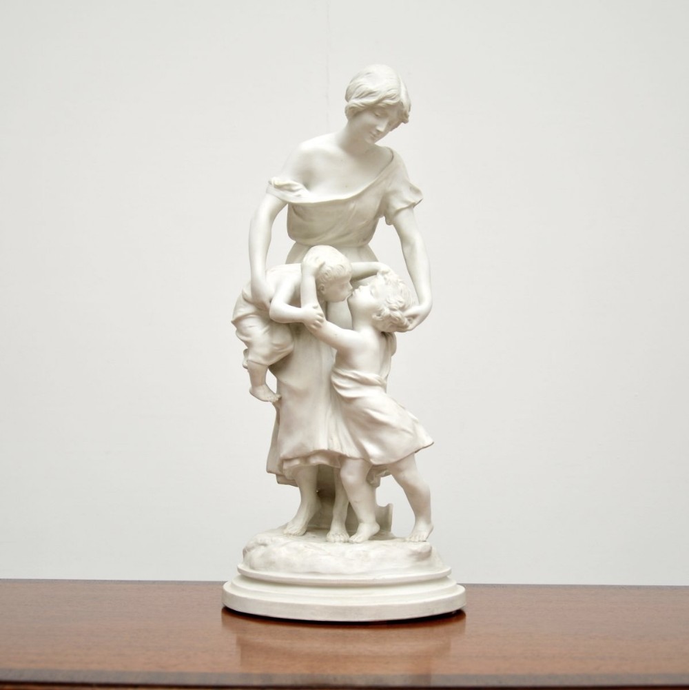 A white bisque porcelain sculpture depicting a standing woman holding and embracing two young children.