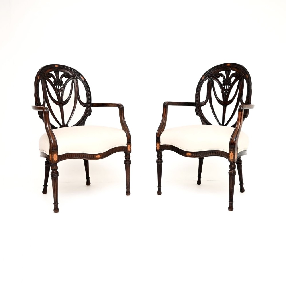 A pair of antique Edwardian mahogany carver armchairs featuring oval openwork backs, satinwood floral inlays, shaped armrests, and off-white upholstered seats.