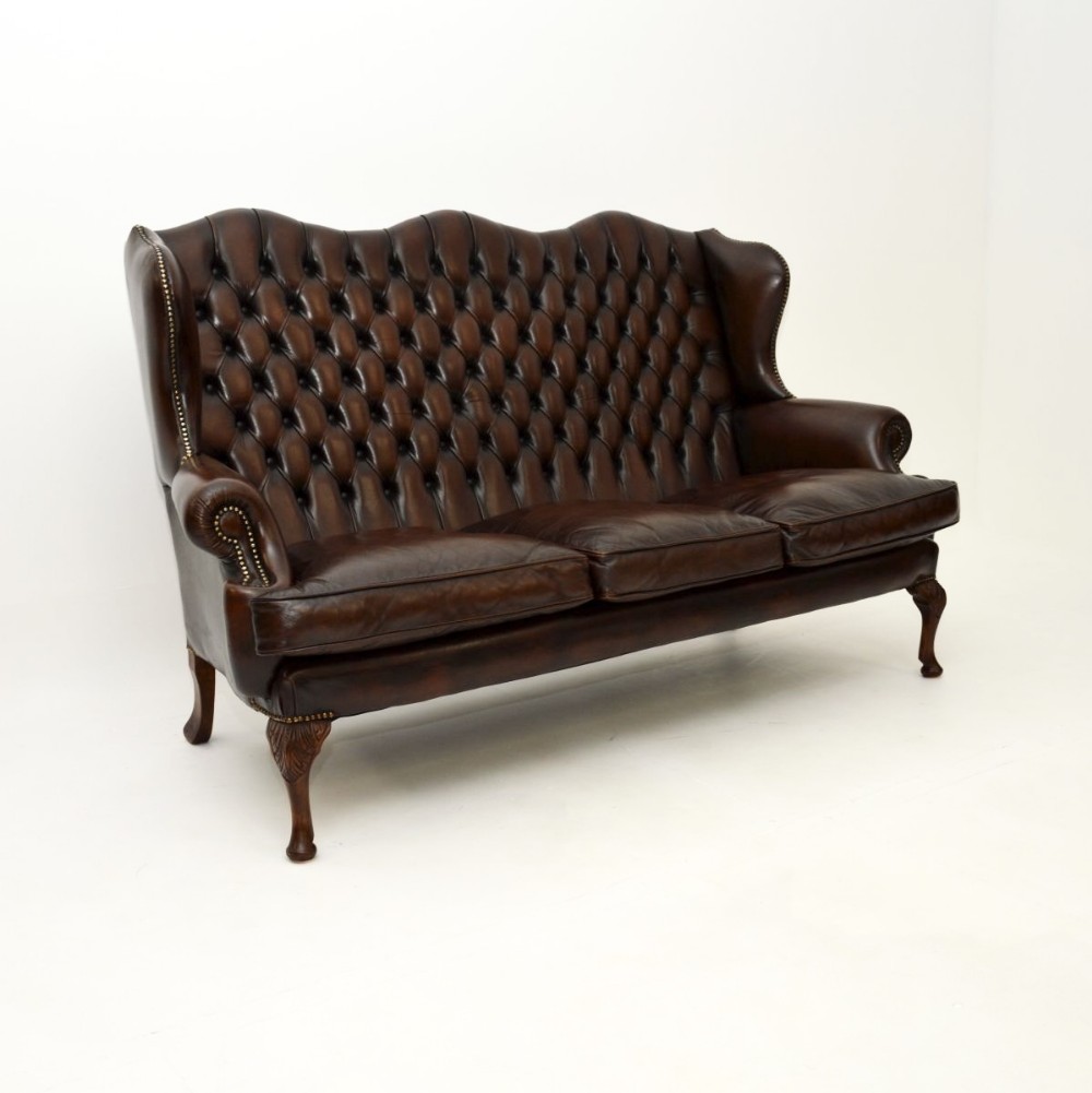 A dark brown leather wingback sofa featuring deep-buttoned tufting, brass stud detailing, loose seat cushions, and carved mahogany cabriole legs.
