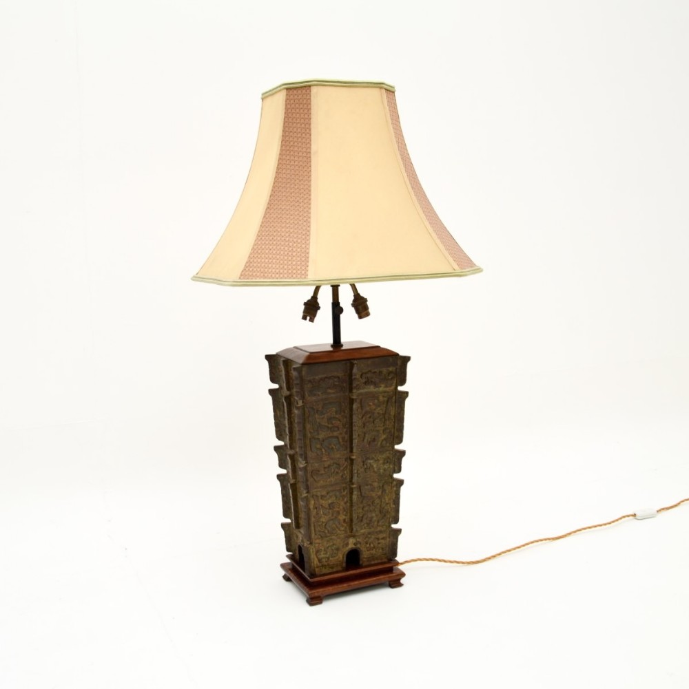 A large patinated bronze table lamp inspired by archaic oriental vessels, mounted on a dark wooden base with a cream fabric lampshade.