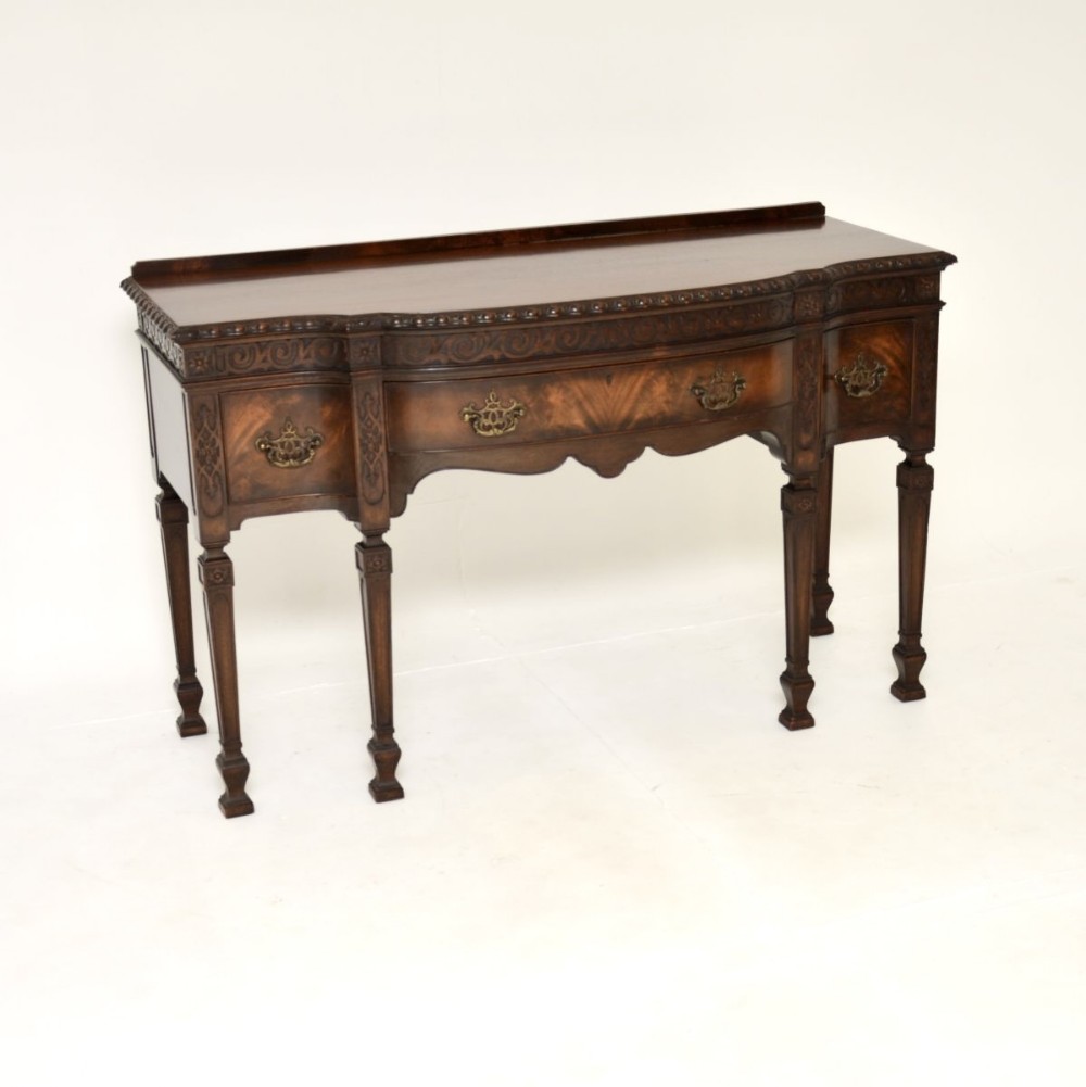 An antique dark mahogany server table featuring a serpentine curved front, carved decorative frieze, three drawers with brass handles, and square tapered legs.