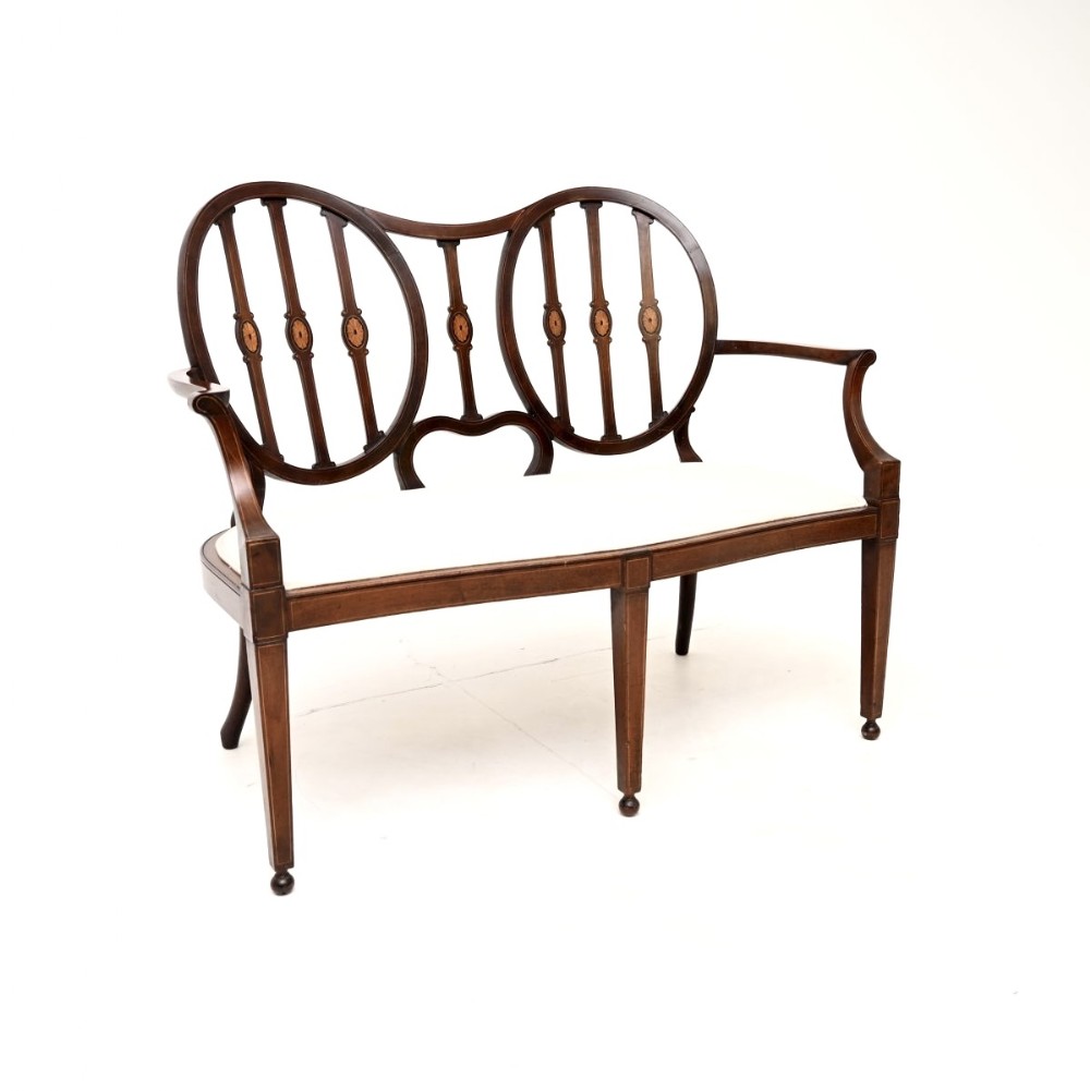 An Edwardian inlaid mahogany settee featuring a double oval backrest with verticle splats, slender curved armrests, tapered legs, and a cream upholstered seat.