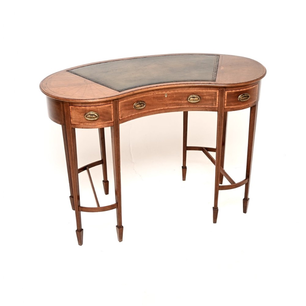 An Edwardian mahogany kidney-shaped desk featuring a dark leather writing surface, satinwood inlay, three drawers with brass handles, and slender tapered legs.