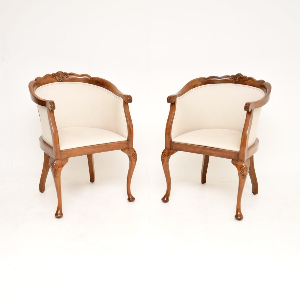 A pair of Queen Anne style walnut tub armchairs featuring cream fabric upholstery, carved cresting rails, and cabriole legs with shell motif details.