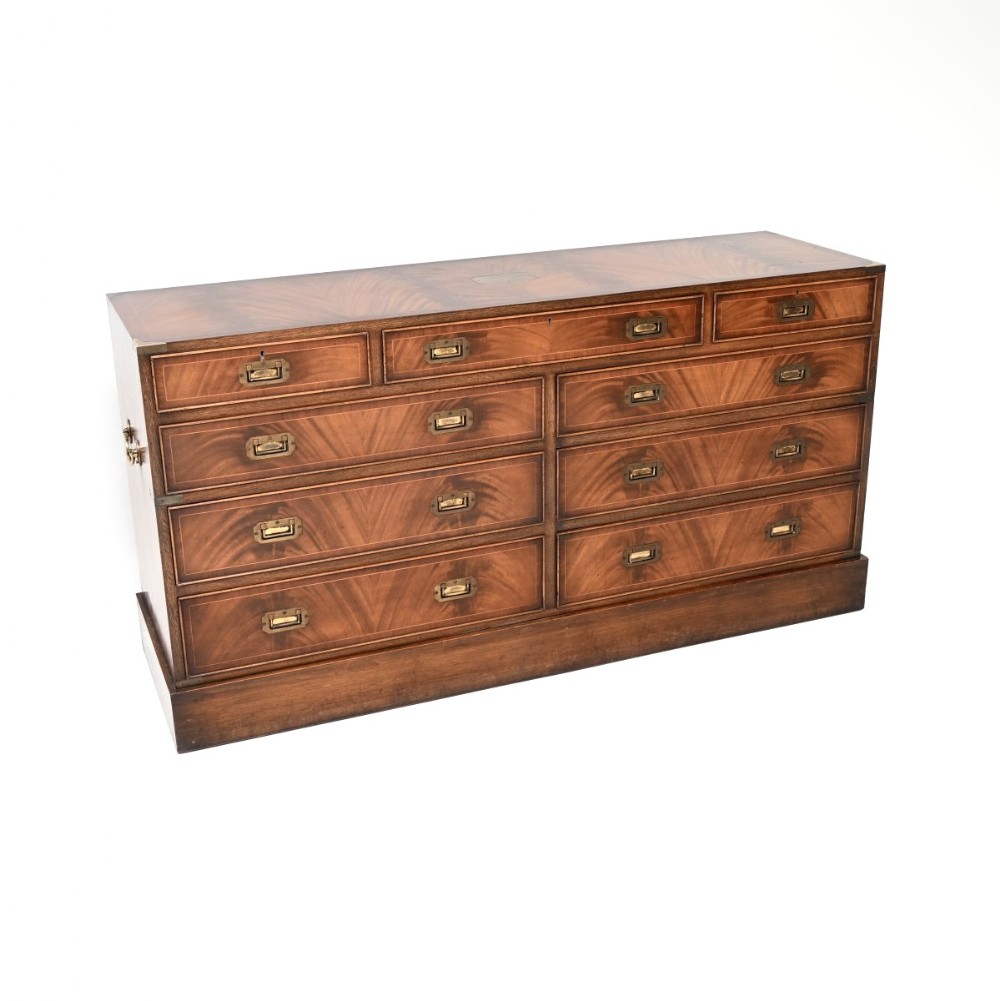 A wide mahogany military campaign style chest of drawers featuring flush recessed brass handles, protective brass corner mounts, and a rich wood grain finish.
