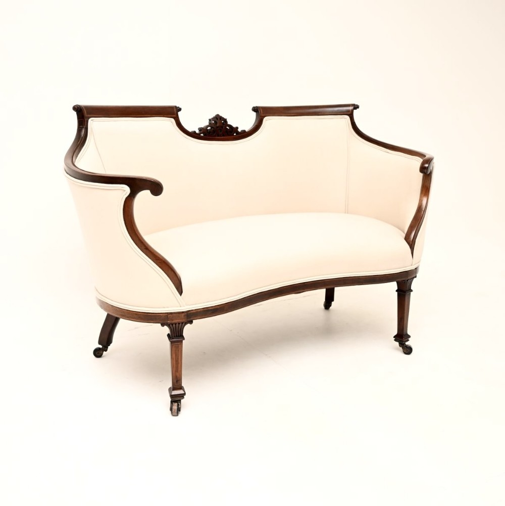 An Edwardian inlaid mahogany settee with light cream upholstery, carved back rail detail, and tapered legs fitted with brass castors.