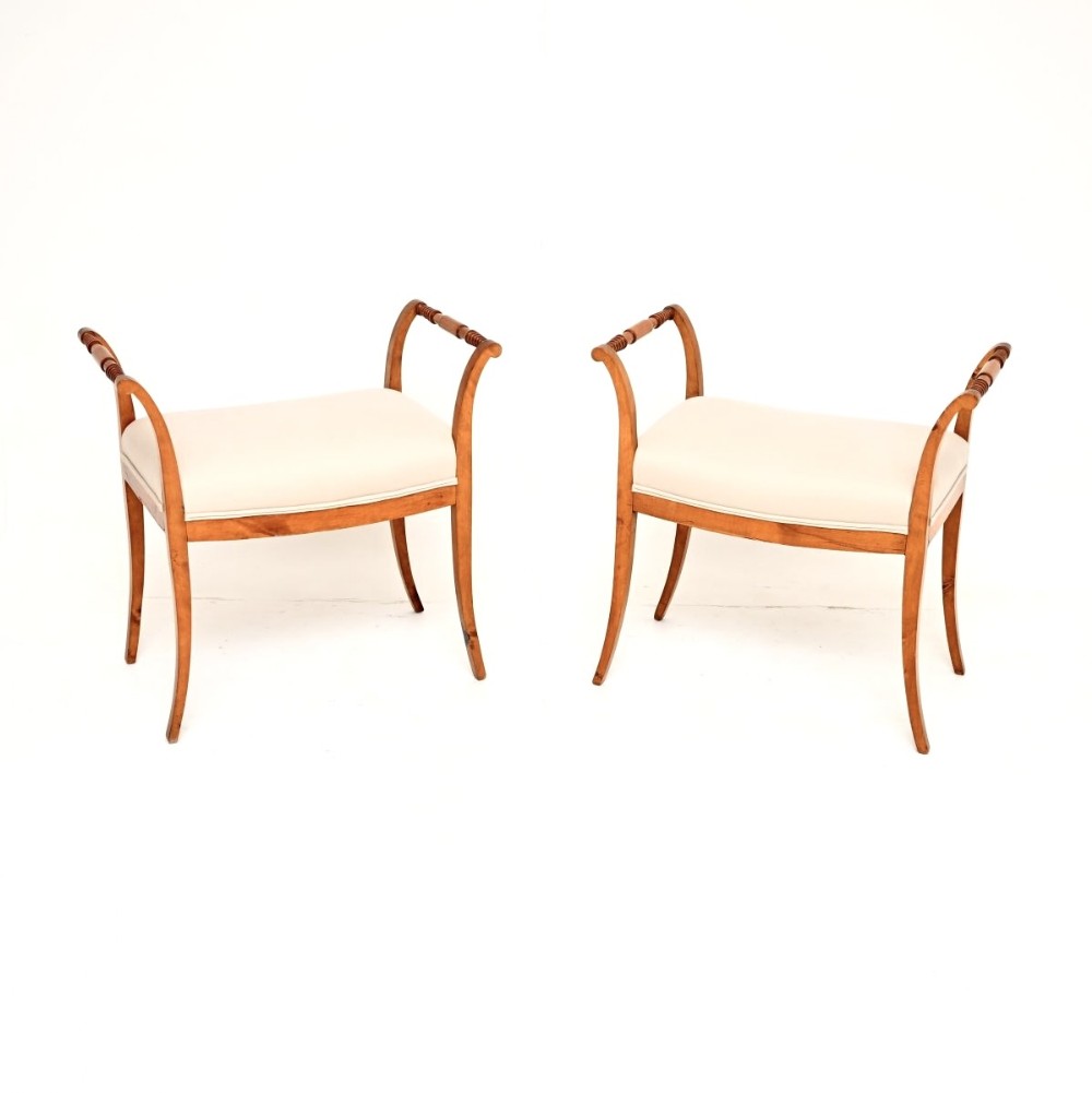 A pair of antique Swedish Biedermeier stools crafted from golden satin birch with swept legs, turned arm handles, and cream upholstered seats.