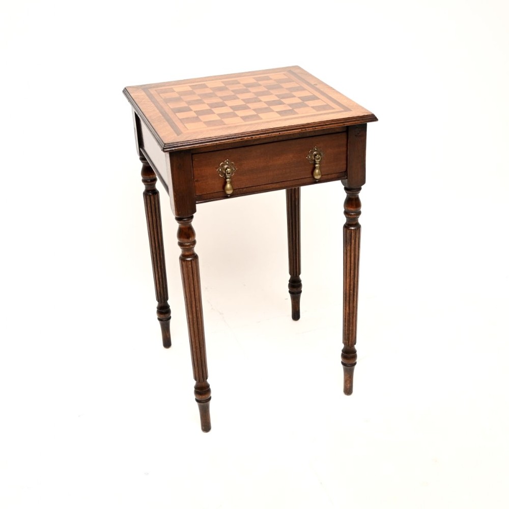 An antique dark wood side table featuring an inlaid checkered game board on the top surface, turned reeded legs, and a single drawer with brass drop handles.