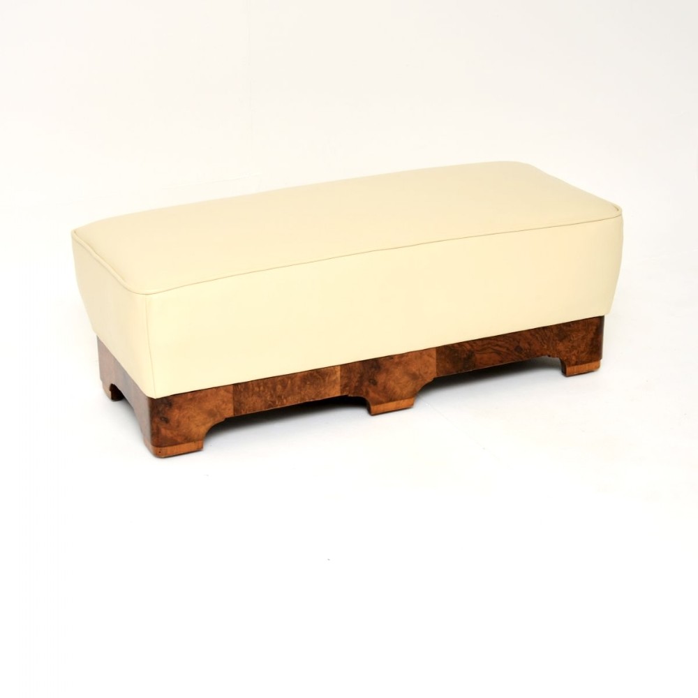 An Art Deco low rectangular stool featuring a cream leather upholstered cushioned top set above a burr walnut plinth base with shaped feet.