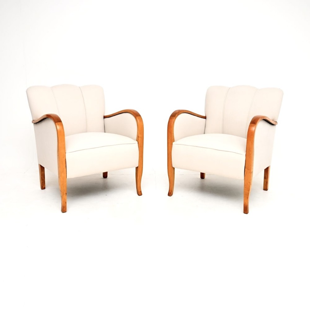 A pair of Swedish Art Deco armchairs featuring off-white fabric upholstery, fluted backrests, and curved satin birch armrests extending down to tapered legs.