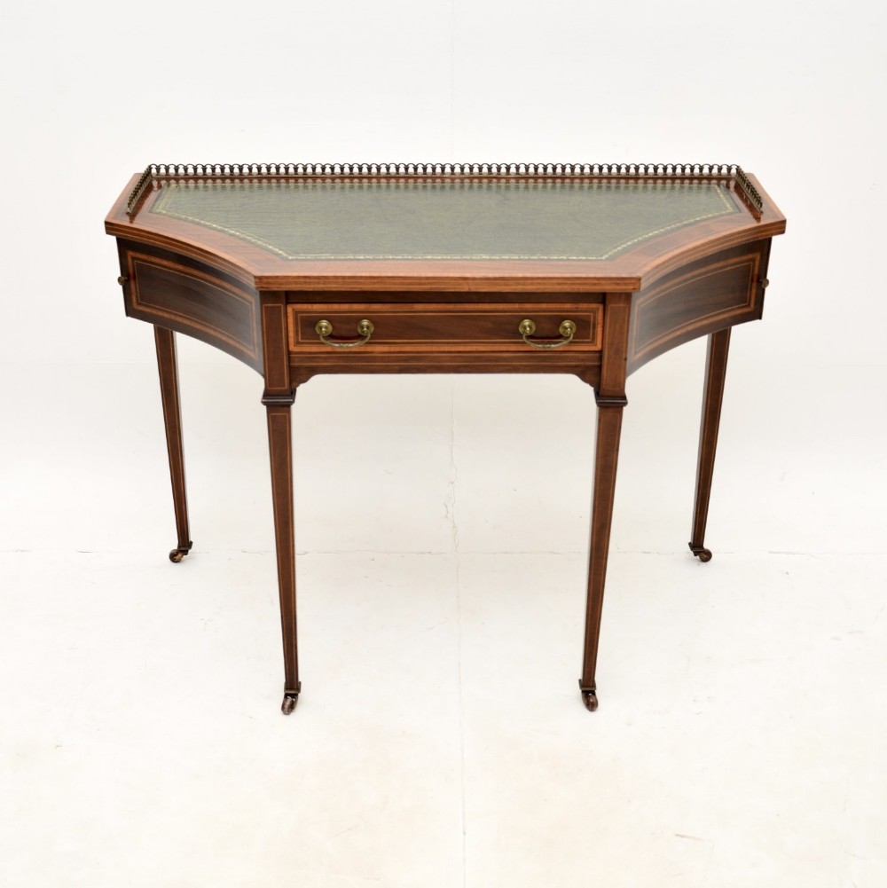 An antique Edwardian mahogany writing table featuring a crescent shape, a green leather top, a brass gallery, and satinwood inlay on tapered legs.