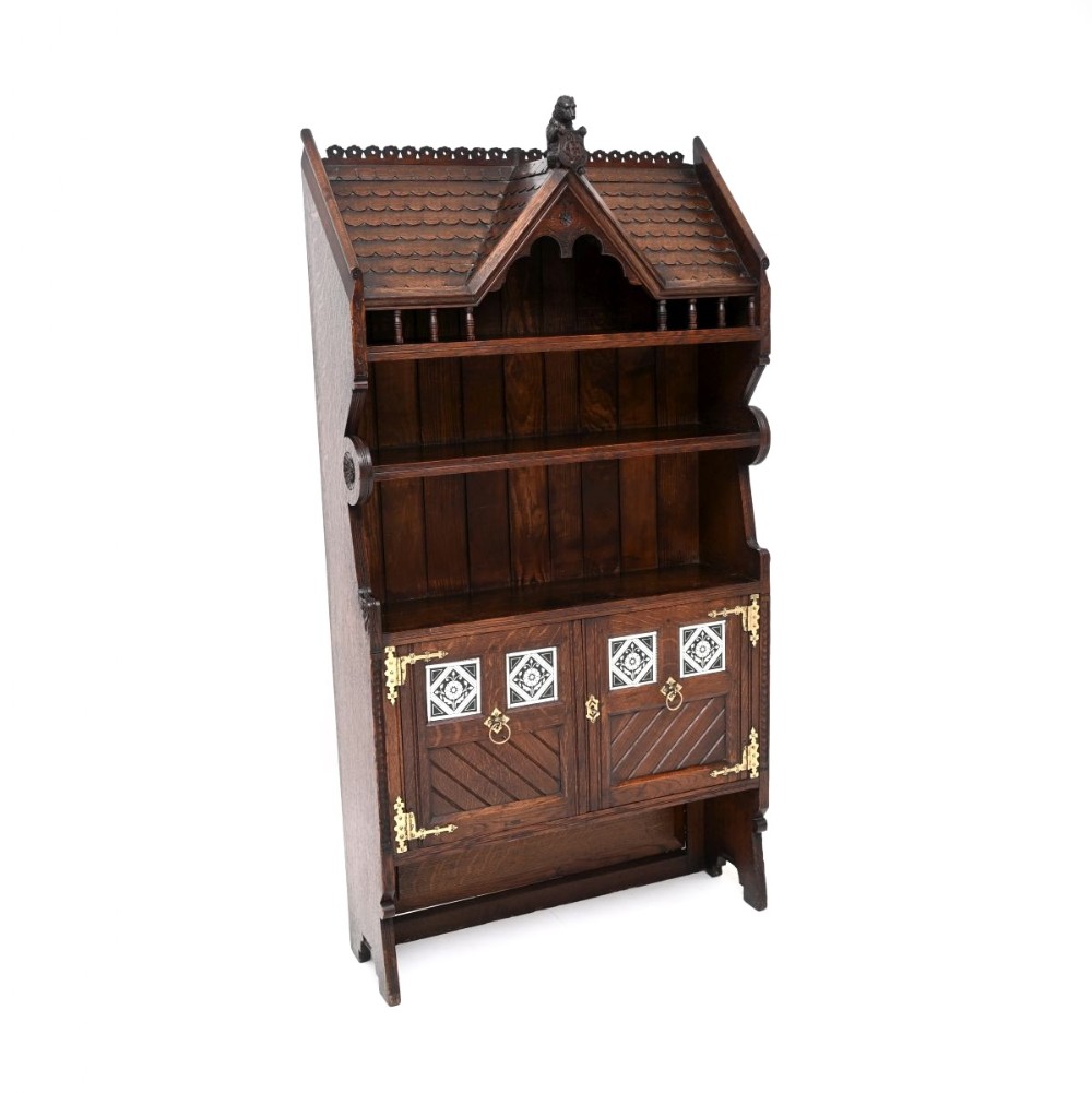 A dark carved oak Aesthetic Movement cabinet featuring a pitched roof canopy, open shelves, and cupboard doors decorated with black and white ceramic tiles and brass hardware.